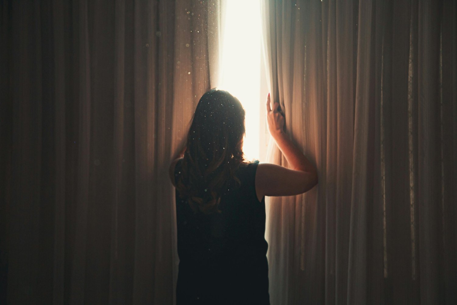 A woman stands by her window in the morning light, gazing outside with a worried expression as intrusive thoughts run through her mind.