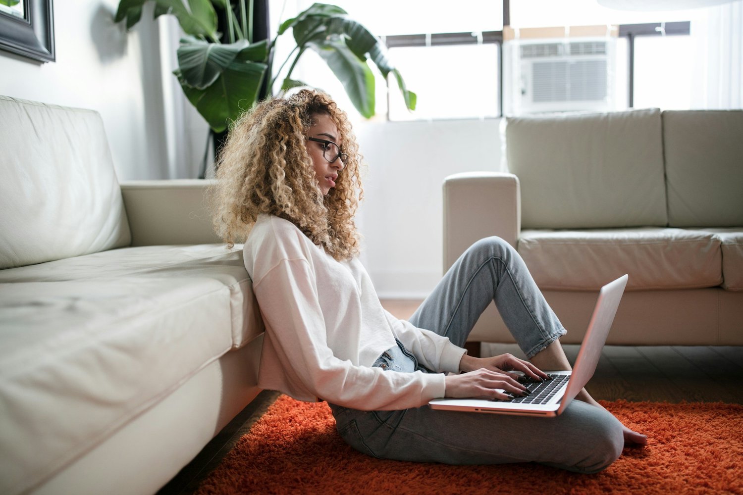 Woman using a laptop while sitting beside a couch in a home setting.