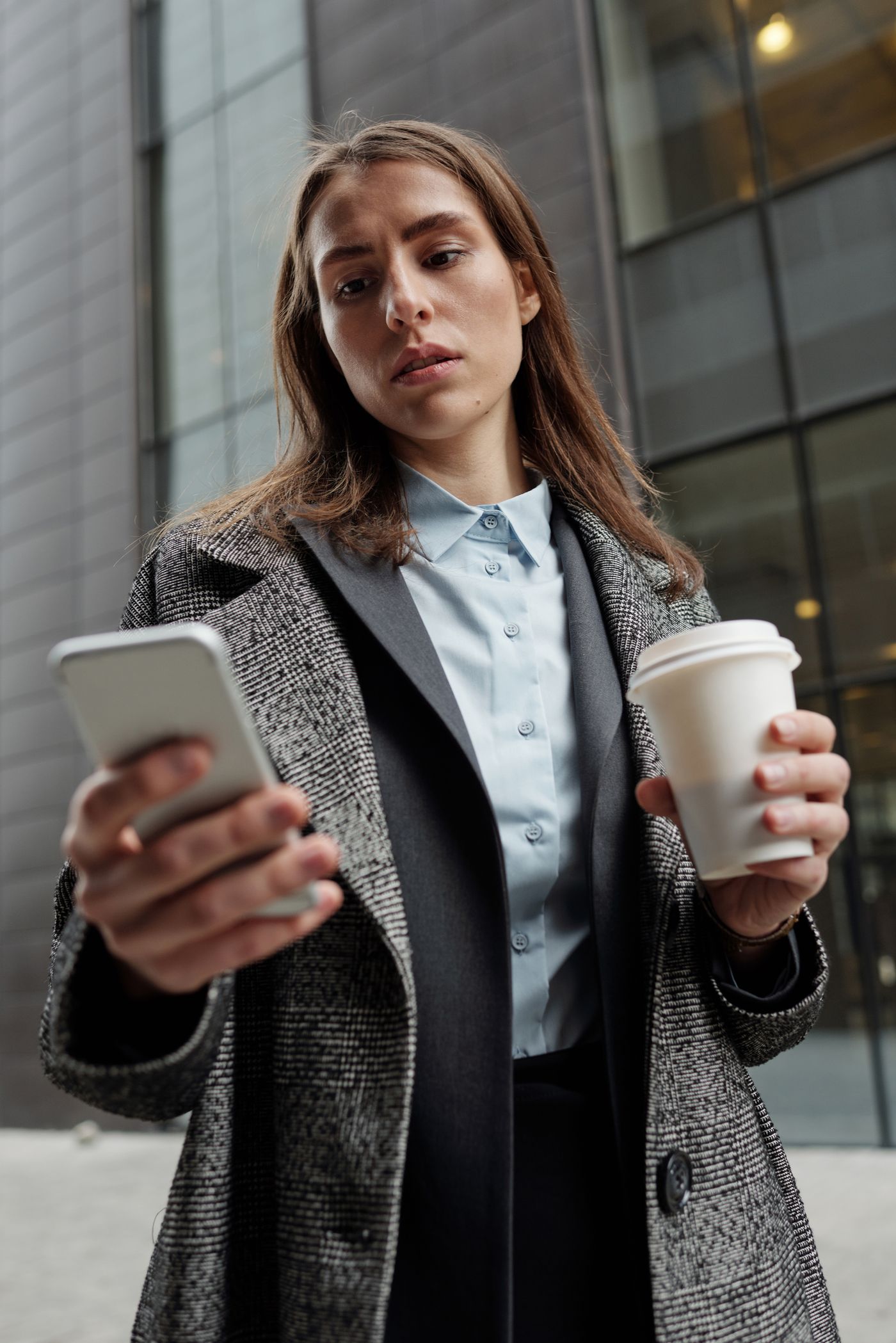 A woman holding a coffee cup while looking at her smartphone in a city setting.