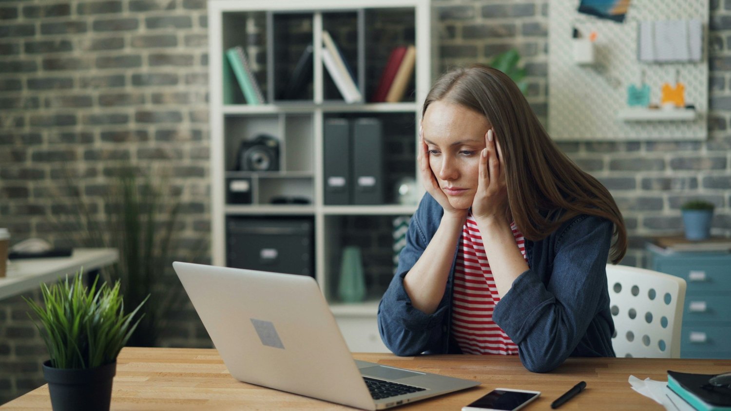 A woman sitting at her laptop with hands on her face, looking overwhelmed and stressed, as if struggling to focus on work.